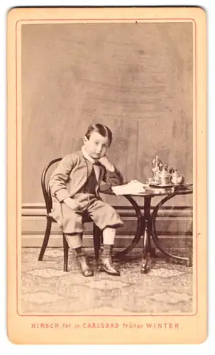 Fotografie Martin Hirsch, Carlsbad, Portrait junger Knabe im Anzug sitzend am Kindertisch, 1873