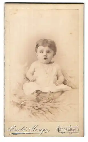 Fotografie Arnold Mange, Rorschach, Kronenstrasse, Baby im weissen Kleidchen auf Felldecke sitzend