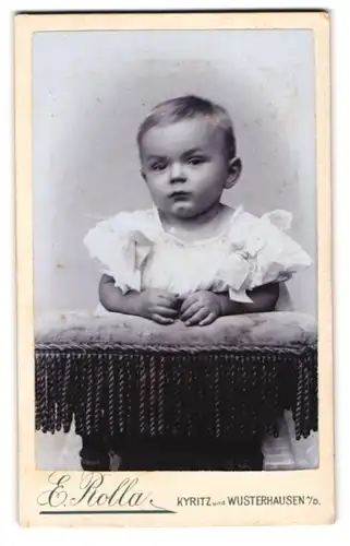 Fotografie E. Rolla, Kyritz, Portrait blondes niedliches Kleinkind im weissen gerüschten Kleidchen