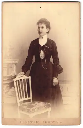 Fotografie L. Haase & Co., Frankfurt, Regierungsstrasse 12, Junge Frau im schwarzen Kleid lehnt an Stuhl