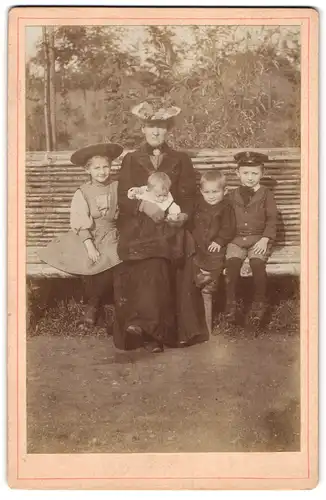 Fotografie unbekannter Fotograf und Ort, Griesgrämig blickende Frau mit ihren Kindern auf einer Parkbank, Mutterglück