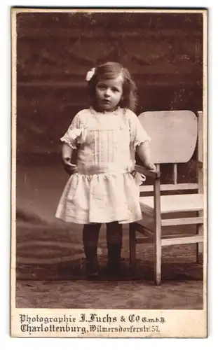 Fotografie J. Fuchs, Berlin, Wilmersdorferstr. 57, Junges Mädchen in weissem Kleid an Stuhl haltend