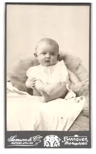 Fotografie Samson & Co., Hannover, Ernst-August-Platz 5, Portrait süsses Baby im weissen Kleidchen auf Fell sitzend