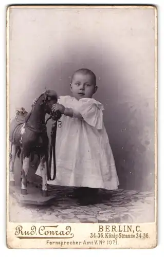 Fotografie Rud. Conrad, Berlin, Königstr. 34-36, Portrait Kleinkind im weissen Kleidchen mit grossem Schaukelpferd