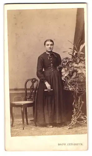 Fotografie Gebr. Bopp, Innsbruck, Portrait Dame im dunklen Biedermeierkleid stehend neben Pflanzengestell