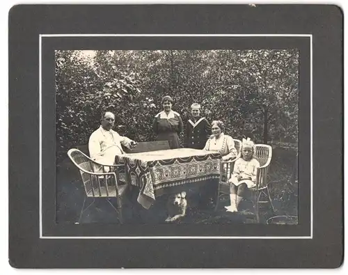 Fotografie unbekannter Fotograf und Ort, Familie mit zwei Kindern im Garten am Tisch, Hund unter dem Tisch