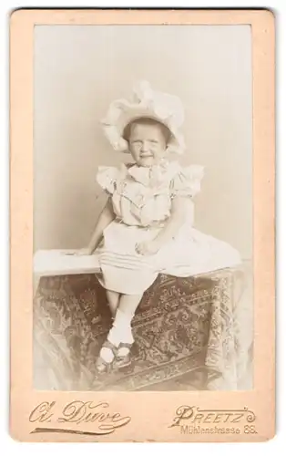 Fotografie A. Duve, Preetz, Mühlenstr. 88, Portrait kleines Mädchen im weissen Kleid mit Haube sitzt auf dem Tisch