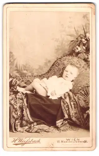 Fotografie H. Weissbach, Wien, niedliches Kleinkind im weissen Kleidchen liegt auf einer Couch