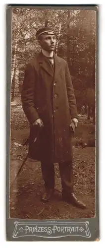 Fotografie unbekannter Fotograf und Ort, Portrait Student im Mantel mit Stock und Schirmmütze im Wald
