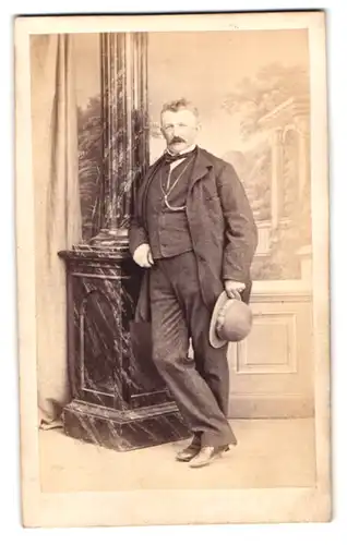 Fotografie Theodor Schlüter, Pinneberg, rundlicher älterer Herr im Anzug mit Melone und Walrossbart lehnt an Mamorsäule