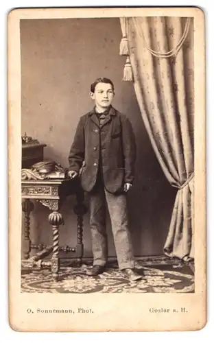 Fotografie O. Sonnemann, Goslar a. H., junger Knabe im Anzug mit Fliege stehend am Sekretär