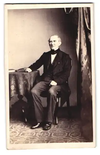 Fotografie F. König, Hamburg, Adolphsplatz 7, älterer Herr im Anzug mit Fliege posiert sitzend im Atelier