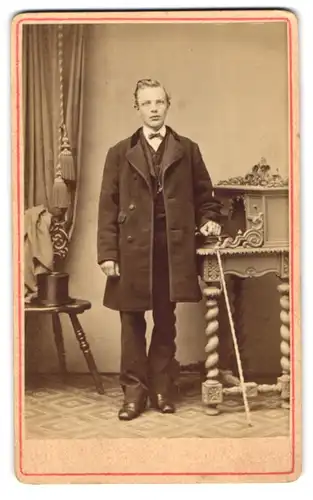 Fotografie unbekannter Fotograf und Ort, junger Herr im Anzug mit Flanierstock und Zylinder im Atelier am Sekretär 