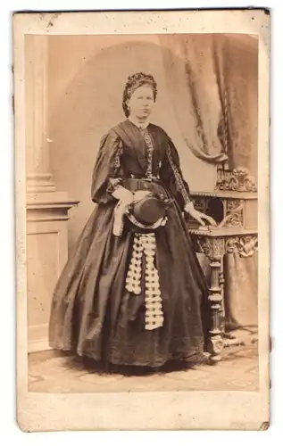 Fotografie M. Kriegsmann, Flensburg, Nordermarkt 49, Dame im Biedermeierkleid mit Damenhut in der Hand am Sekretär