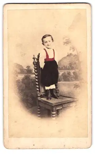 Fotografie H. Strube, Löbau, Bahnhofstr. 316, niedlicher junger Knabe in Latzhosde steht auf Stuhl, Hand Koloriert