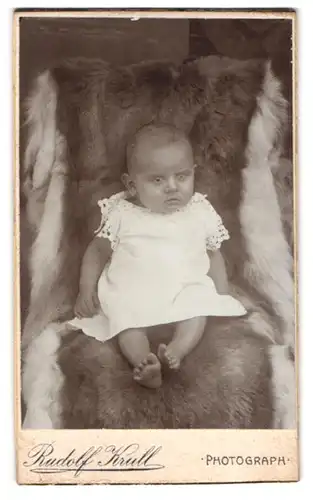 Fotografie Rudolf Krull, Ort unbekannt, Süsses Kleinkind im weissen Hemd sitzt auf Fell