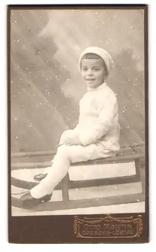 Fotografie Otto Martin, Dresden, Portrait junger Knabe in Winterkleidung mit Strumpfhose auf seinem Schlitten sitzend