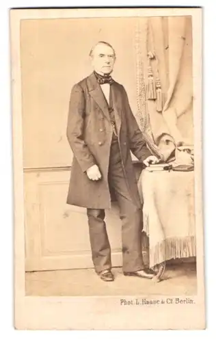 Fotografie L. Haase & Co., Berlin, Grosse Friedrichstr. 178, älterer Herr im Anzug mit Fliege stehend am Tisch