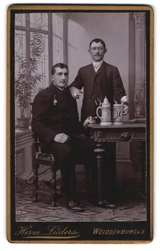 Fotografie Herm. Lüders, Weissenburg a. S., Portrait zwei Herren in Anzügen mit Bierkrügen auf dem Tisch