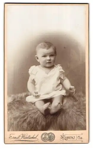 Fotografie Ernst Hentschel, Kamenz i. Sa., Hoyerswerdaerstr. 30, Portrait süsses Baby im weissen Hemdchen auf Fell sitzend