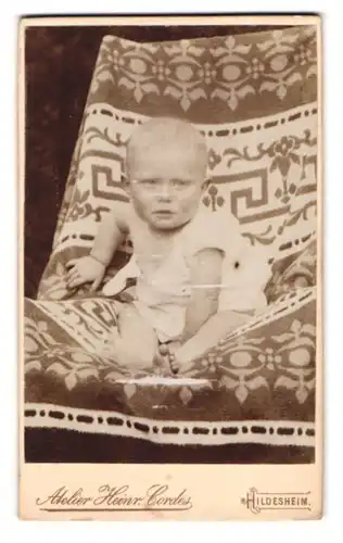 Fotografie Heinr. Cordes, Hildesheim, Zingel 36, Portrait kleiner Bube im weissen Hemdchen auf einer bedruckten Decke