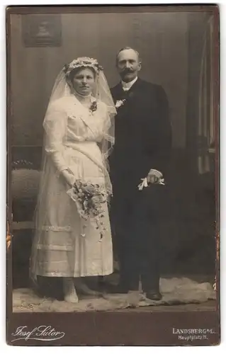 Fotografie Josef Sutor, Landsberg a. L., Hauptplatz 11, Eheleute im Hochzeitskleid und Anzug mit Brautstrauss