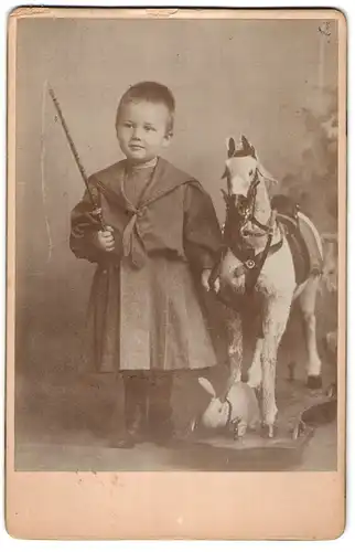 Fotografie unbekannter Fotograf und Ort, Portrait Kleinkind im Kleidchen mit Schaukelpferd und Plüschhasen, Peitsche