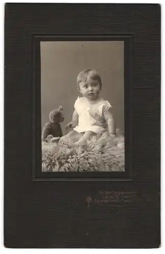 Fotografie Fr. Bruggemann, Leipzig, Eisenbahnstr. 1, Kleinkind im Kleidchen mit seinem Teddybär auf einem Fell sitzend