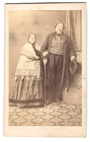 Fotografie unbekannter Fotograf und Ort, Portrait korpulentes Paar im Kleid mit Decke und Anzug mit Stock und Hut, 1868