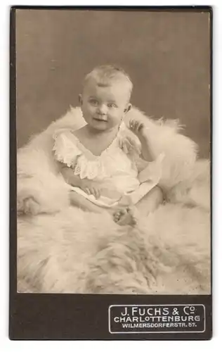 Fotografie J. Fuchs & Co., Berlin-Charlottenburg, Wilmersdorferstr. 57, Süsses Kleinkind im Hemd sitzt auf Fell
