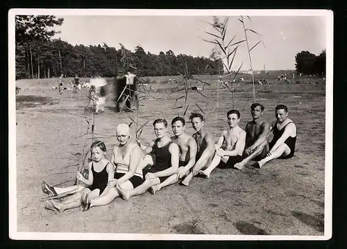 Fotografie Burschen & Frauen in Badebekleidung hintereinader am Strand sitzend, Schnappschuss