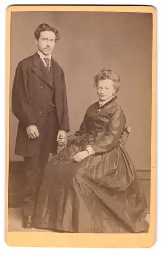 Fotografie M. Wild, Ort unbekannt, Portrait junges Paar im seidenen Biedermeierkleid ind Anzug halten Händchen