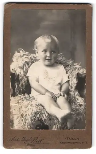 Fotografie Arth. Josef Bett, Tulln, Kaiser-Wilhelm-Str. 19, Portrait süsses blondes Kleinkind auf Fell sitzend