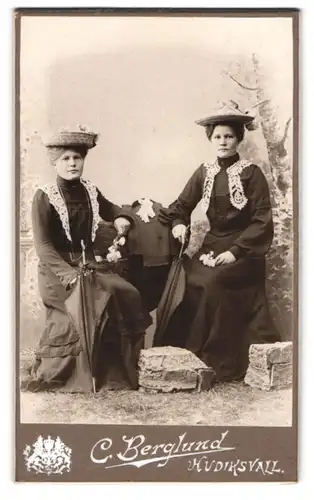 Fotografie C. Berglund, Hudiksvall, Portrait zwei Damen in dunklen Kleidern mit Spitzenbesatz und breiten Hüten, Schirm