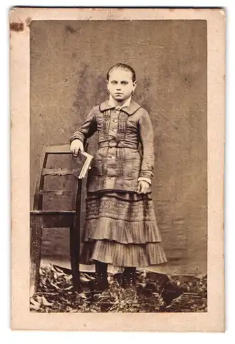 Fotografie unbekannter Fotograf und Ort, niedliches Mädchen im karierten Kleid posiert neben einem Stuhl