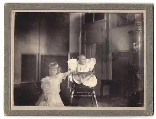 Fotografie unbekannter Fotograf und Ort, zwei niedliche kleine Kinder im Wohnzimmer mit altem Heizofen und Kinderstuhl