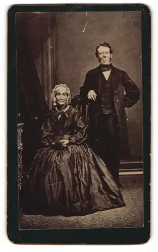 Fotografie unbekannter Fotograf und Ort, Portrait älteres Paar im seidenen Biedermeierkleid und Anzug posiert im Atelier