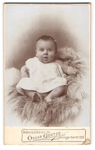Fotografie Oskar Goetze, Königsberg i. Pr., Weissgerberstr. 229, Portrait süsses Baby im Kleidchen auf Fell sitzend