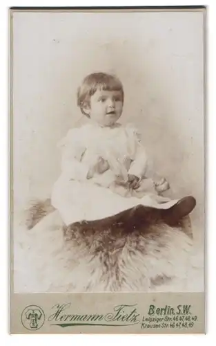 Fotografie Hermann Tietz, Berlin-SW, Leipziger-Str. 46-49, Süsses Kleinkind im Kleid mit Puppe sitzt auf Fell