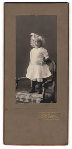 Fotografie A. Grosskopf, Aalen, Kleines Mädchen mit Locken und Haarschleife, auf einem Sessel stehend