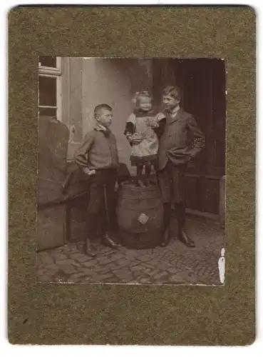 Fotografie unbekannter Fotograf und Ort, Zwei Jungen mit lachendem kleinem Mädchen auf einer Tonne
