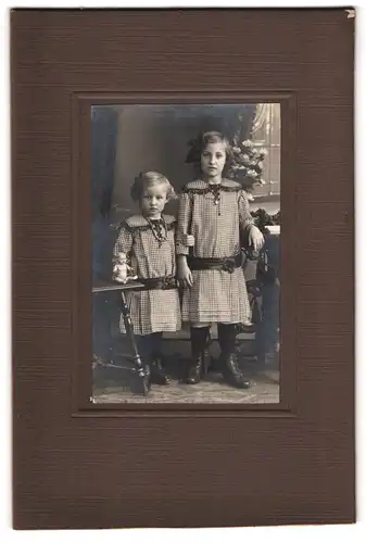 Fotografie unbekannter Fotograf und Ort, Portrait zwei niedliche Schwestern in karierten Kleidern m. Puppe auf dem Tisch