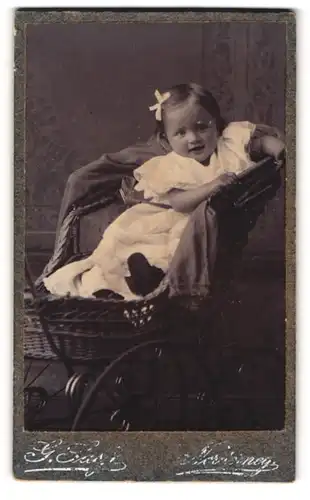 Fotografie G. Sass, Norderney, Portrait süsses Mädchen im weissen Kleidchen sitzt im Kinderwagen mit Haarschleife