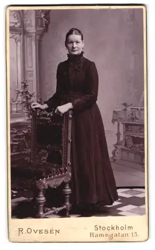 Fotografie R. Oversen, Stockholm, Hamngatan 15, ältere Dame im dunklen Kleid am Gründerzeitstuhl stehend