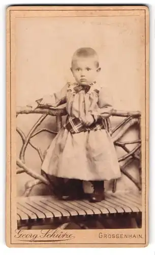 Fotografie Georg Schütze, Grossenhain, Johannes-Allee 64b, Portrait süsses Kleinkind im Kleidchen auf einer Bank stehend