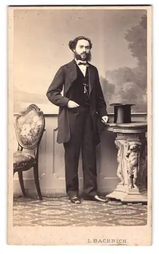 Fotografie L. Bachrich, Wien, Portrait junger Mann im Anzug mit buschigem Backenbart und Zylinder auf dem Tisch