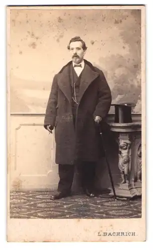 Fotografie L. Bachrich, Wien, Herr im Anzug mit Wintermantel, Zylinder, Flanierstock, rauchend im Atelier