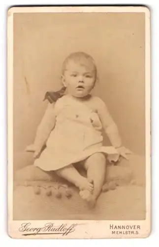 Fotografie Georg Rudloff, Hannover, Mehlstrasse 5, Baby mit offenem Mund und übereinandergelegten Füssen auf einem Kissen