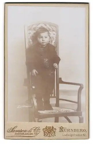 Fotografie Samson & Co., Nürnberg, Ludwigstrasse 42, Ernst blickender Junge im Sonntagsstaat, auf einem Stuhl stehend