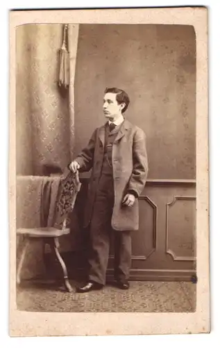 Fotografie unbekannter Fotograf und Ort, Portrait junger Mann im grauen Anzug stehend an einem Stuhl